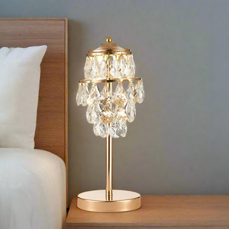This table lamp features a gold base with a slender stem, topped by cascading crystal pendants arranged in tiers. The design combines structured elegance with a luxurious, chandelier-like aesthetic.