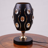 A black globe table lamp with a geometric design, adorned with crystal beads, gold accents, and a sleek base, sit on a wooden table. It features an LED light with an E27 bulb, casting a warm, stylish glow.