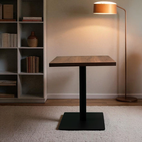 Modern table with black base and wooden top in a room with a bookshelf and floor lamp.
