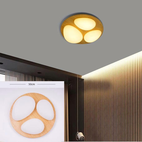 A circular flush-mount ceiling light with three teardrop-shaped white light zones inside a light wood frame, installed flat on a grey ceiling above wood-paneled walls, giving off warm diffused light.

