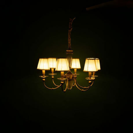 "A golden chandelier, softly illuminated in a dimly lit room, features multiple gracefully curved arms. Each arm holds a white pleated lampshade with intricate decorative trim. At the centre of the chandelier is a beautiful glass column, and it is suspended from a gold-colored rod, adding a touch of elegance to the room's atmosphere.