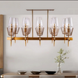 A sleek gold chandelier with a geometric design and multiple glass-shaded lights hangs from a light-colored ceiling, illuminating a sophisticated dining room. Below, an elegant brown dining table is paired with white chairs, and the table is set with a decorative vase and neatly arranged plates