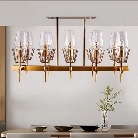 A sleek gold chandelier with a geometric design and multiple glass-shaded lights hangs from a light-colored ceiling, illuminating a sophisticated dining room. Below, an elegant brown dining table is paired with white chairs, and the table is set with a decorative vase and neatly arranged plates