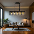 A modern rectangular chandelier with five exposed vintage-style light bulbs enclosed in glass shades, suspended by two twisted black rods. The fixture hangs above a cozy, minimalist living room featuring a beige sectional sofa, a neutral area rug, wooden flooring, and a potted plant in the corner.









