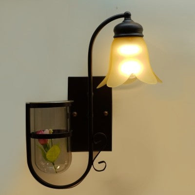 A rustic-inspired wall light with a brown metal  frame, a curved arm, and a frosted glass shade in a flower-like shape equipped with a led lamp E27. Below it, a small decorative glass shelf holds a flower.
