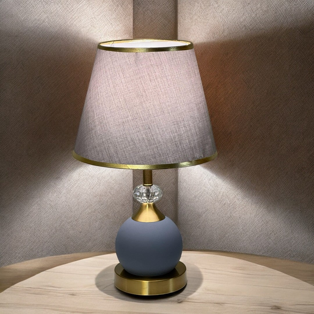 Minimalist round table lamp with a grey fabric shade and matte blue base, placed on a wooden table against a soft-lit beige wall.
