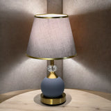 Minimalist round table lamp with a grey fabric shade and matte blue base, placed on a wooden table against a soft-lit beige wall.
