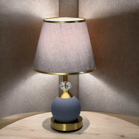 Minimalist round table lamp with a grey fabric shade and matte blue base, placed on a wooden table against a soft-lit beige wall.
