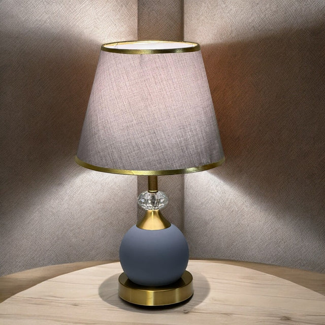 Minimalist round table lamp with a grey fabric shade and matte blue base, placed on a wooden table against a soft-lit beige wall.
