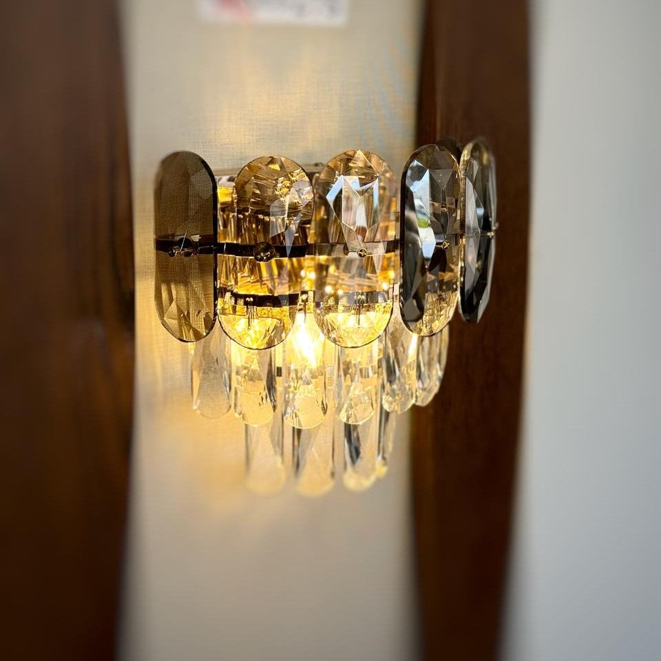 A decorative gold wall light featuring grey circular elements with cascading crystal droplets, reflecting warm light beautifully.