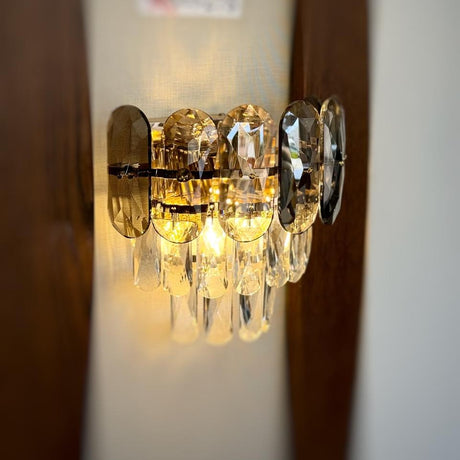 A decorative gold wall light featuring grey circular elements with cascading crystal droplets, reflecting warm light beautifully.
