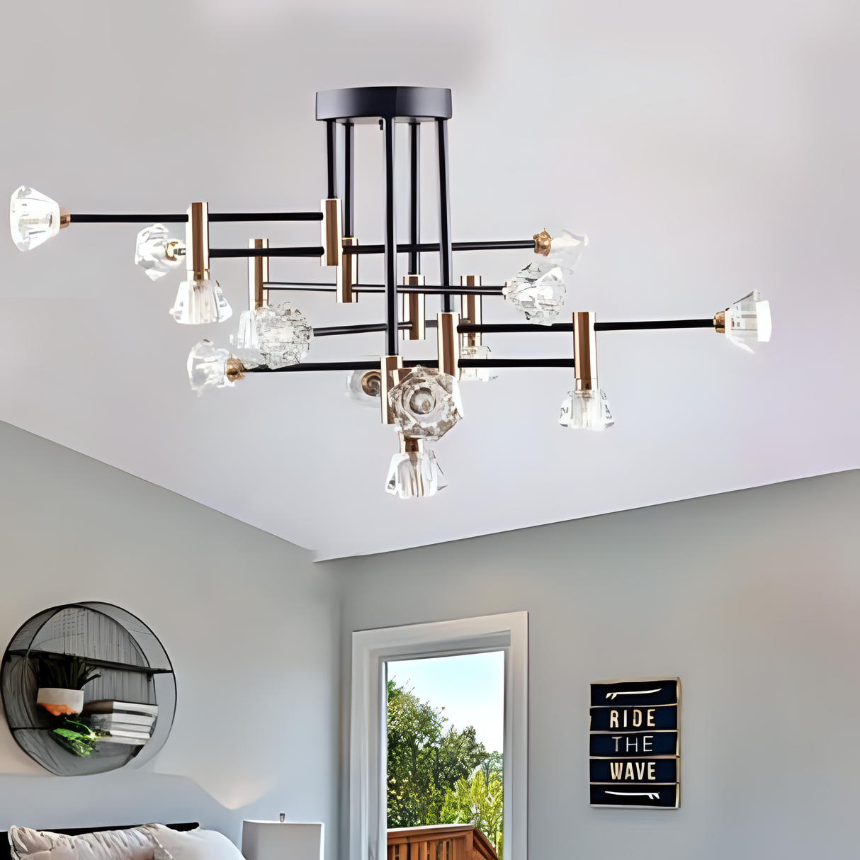 A modern black and gold chandelier with clear, sculpted glass light covers, featuring multiple branching arms in a starburst arrangement, installed on a white ceiling in a stylish living room with minimalist decor, including a round wall shelf, a cozy sofa, and a decorative sign that reads 'Ride The Wave'