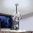 A contemporary circular crystal pendant chandelier with a layered design, featuring clear glass panels and a mirrored frame, hangs from a light-colored ceiling and casts elegant light over a sophisticated dining room. Below, a sleek dining table is paired with white chairs, and a decorative vase serves as the centerpiece.