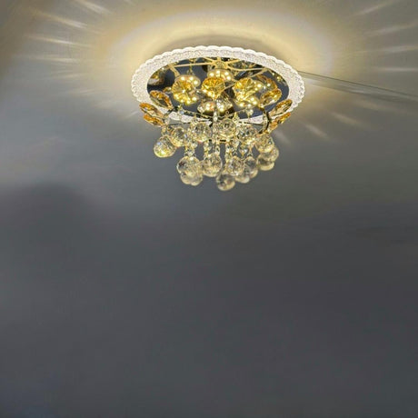 A compact, ceiling-mounted chandelier featuring a gold-toned metal frame and a cluster of teardrop-shaped crystal pendants. The fixture casts warm LED lighting, creating a decorative sunburst reflection on the ceiling. 