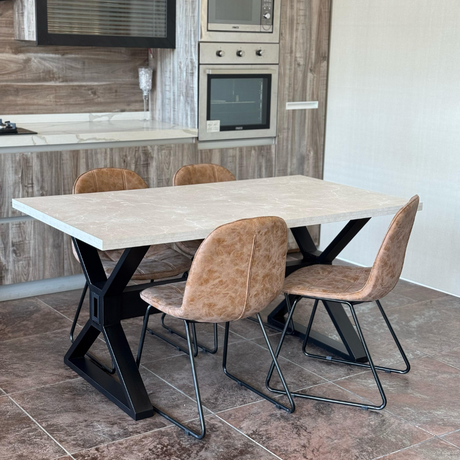 A modern dining set featuring a rectangular wooden-top table with a black metal X-shaped base, paired with four brown faux-leather chairs with metal legs. The set is placed in a contemporary kitchen with wood-paneled walls, built-in stainless steel appliances, and a white countertop