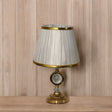 Elegant gold table lamp with a pleated white shade with a gold trim and a crystal accent in the center, placed on a wooden surface against a wood-paneled background.
