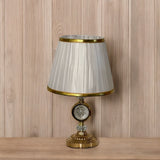Elegant gold table lamp with a pleated white shade with a gold trim and a crystal accent in the center, placed on a wooden surface against a wood-paneled background.