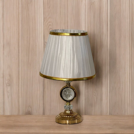 Elegant gold table lamp with a pleated white shade with a gold trim and a crystal accent in the center, placed on a wooden surface against a wood-paneled background.