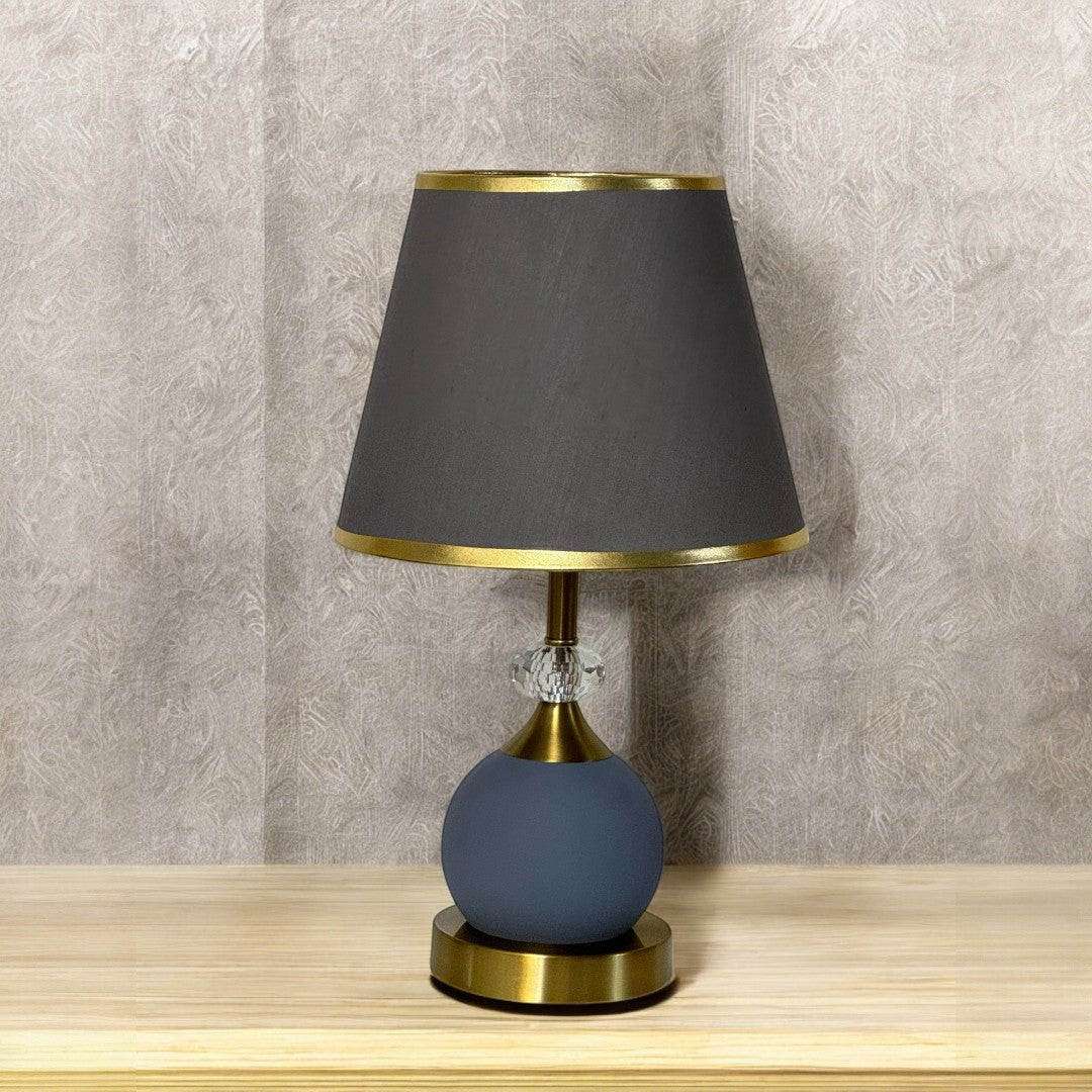 Modern round table lamp with a dark grey shade and matte blue base, placed on a wooden surface against a textured beige wall.

