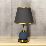 Modern round table lamp with a dark grey shade and matte blue base, placed on a wooden surface against a textured beige wall.
