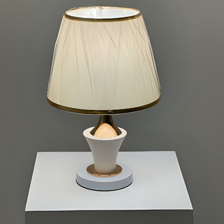 Luxury cream and gold table lamp with a slightly flared pleated lampshade wrapped in protective plastic, placed on a white table.

