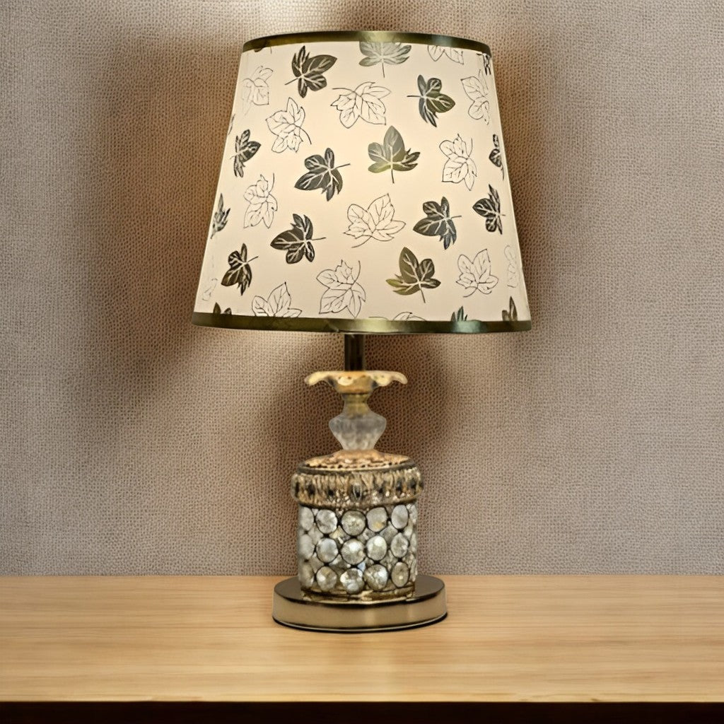 A table lamp with a gold and glass base, and a white fabric lampshade adorned with black floral patterns, set against a beige wall.