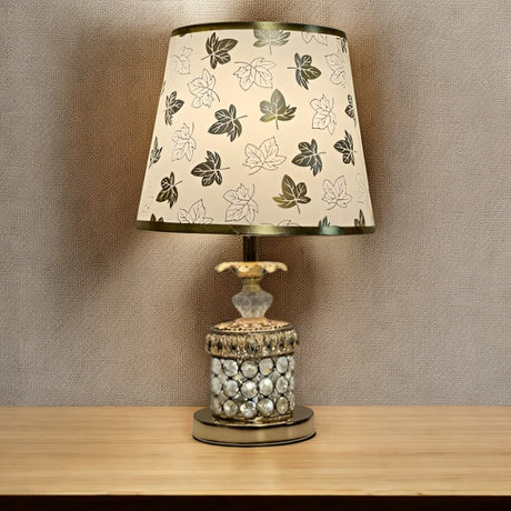 A table lamp with a gold and glass base, and a white fabric lampshade adorned with black floral patterns, set against a beige wall.