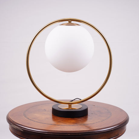 This modern table lamp features a striking circular metal frame that elegantly encases a frosted glass globe, creating a sleek and contemporary aesthetic. Placed on a wooden table. 