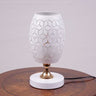 A white modern table lamp with a geometric design, gold accents, and a sleek base sits on a wooden table. It features an LED light with an E27 bulb, casting a warm, stylish glow.