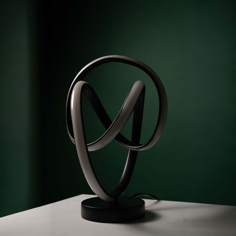 A black matte-finished table lamp with an abstract, curved metal design, placed against a dark green background. The lamp is turned off, showcasing its sculptural form.