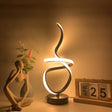 A decorative black table lamp with a unique swirling LED design, placed next to a wooden calendar and a flower vase.
