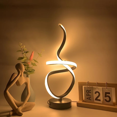 A decorative black table lamp with a unique swirling LED design, placed next to a wooden calendar and a flower vase.
