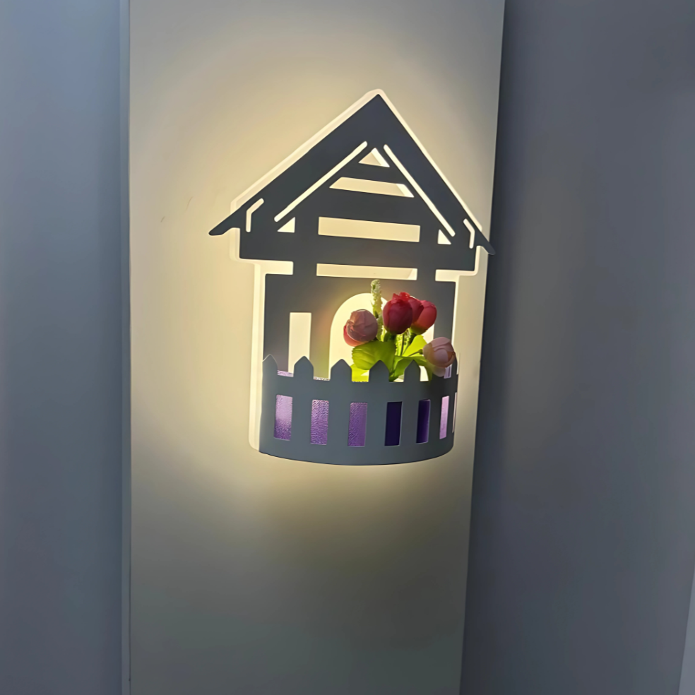 Wall-mounted LED light fixture designed in the shape of a white house , featuring a cork-style built-in compartment holding artificial pink and red tulip flowers. The light has a whimsical, cottage-inspired aesthetic and is mounted on a plain white wall.

