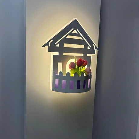 Wall-mounted LED light fixture designed in the shape of a white house , featuring a cork-style built-in compartment holding artificial pink and red tulip flowers. The light has a whimsical, cottage-inspired aesthetic and is mounted on a plain white wall.
