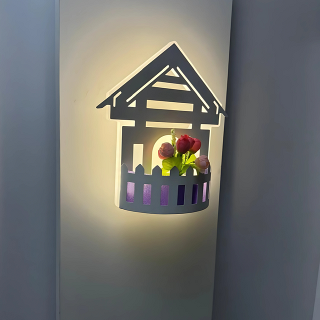 Wall-mounted LED light fixture designed in the shape of a white house , featuring a cork-style built-in compartment holding artificial pink and red tulip flowers. The light has a whimsical, cottage-inspired aesthetic and is mounted on a plain white wall.
