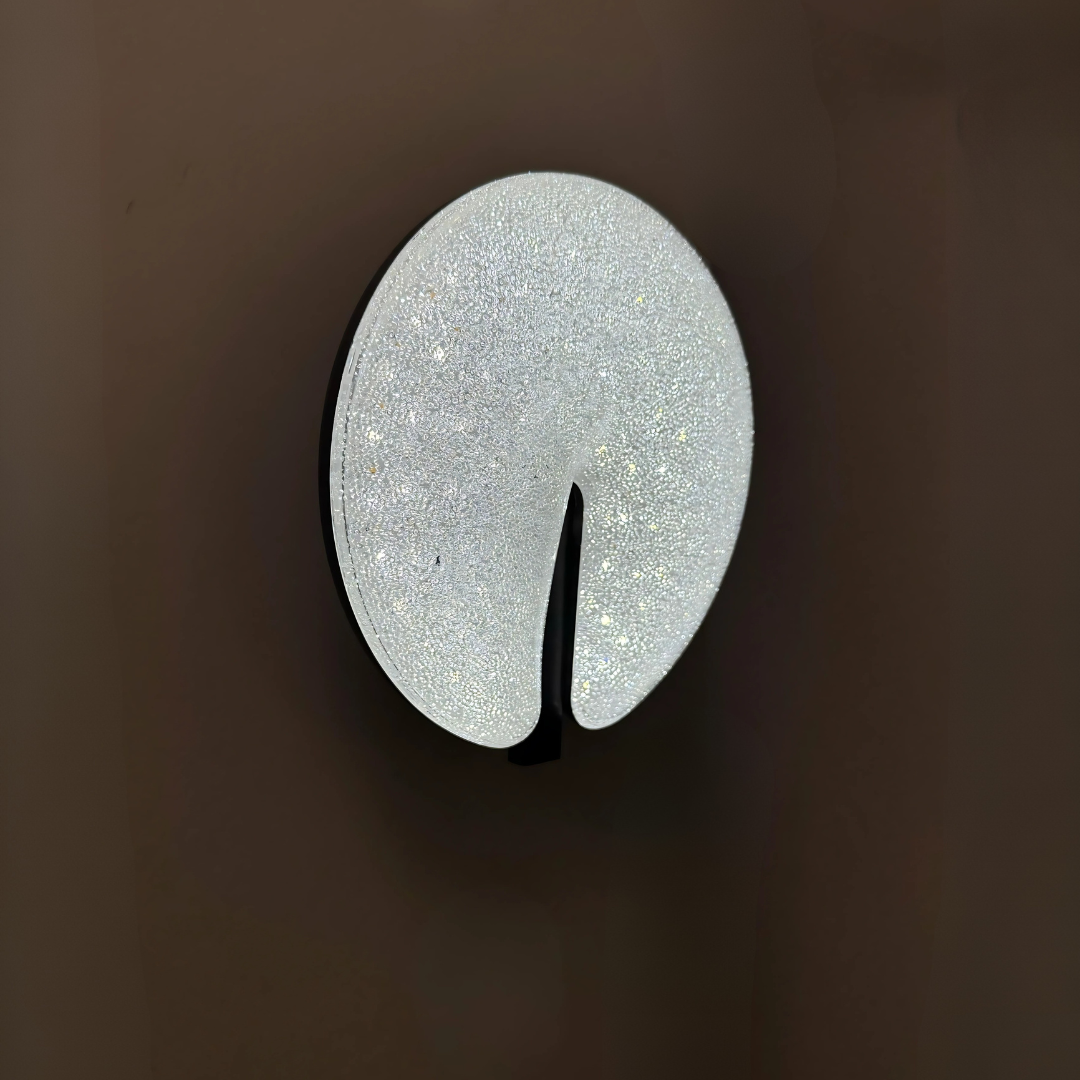 Modern circular LED wall light fixture with a sparkling textured surface and a sleek brown cut-out design, mounted on a dark-colored wall.
