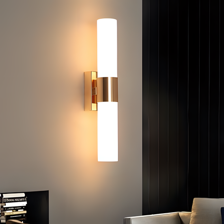 A contemporary gold wall sconce with two cylindrical frosted glass diffusers, mounted on a dark vertical panel.
