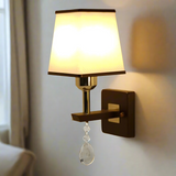 A brown and silver wall light with a rectangular fabric shade and a hanging crystal ornament, adding a touch of elegance turned on, casting warm light.