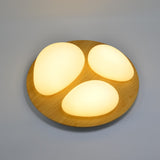 A circular flush-mount ceiling light with three teardrop-shaped white light zones inside a light wood frame, installed flat on a grey ceiling .
