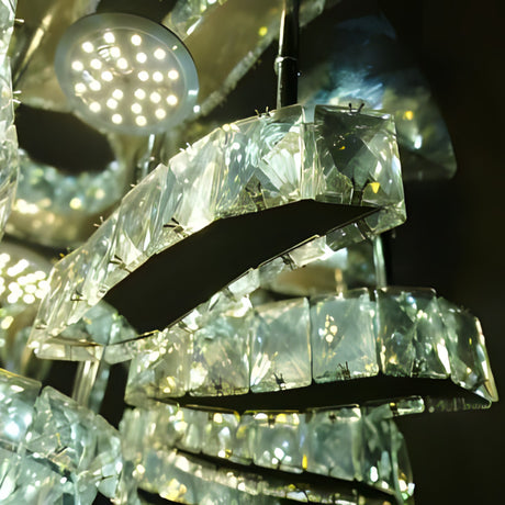A luxurious crystal chandelier with curved, overlapping rectangular glass segments, suspended from the ceiling in a dark space; it reflects bright white light in multiple directions.

