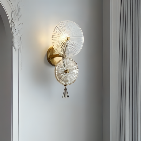 Elegant wall sconce featuring two overlapping circular glass shades with radial etched patterns, mounted on a gold base against a light gray wall with classic molding details.