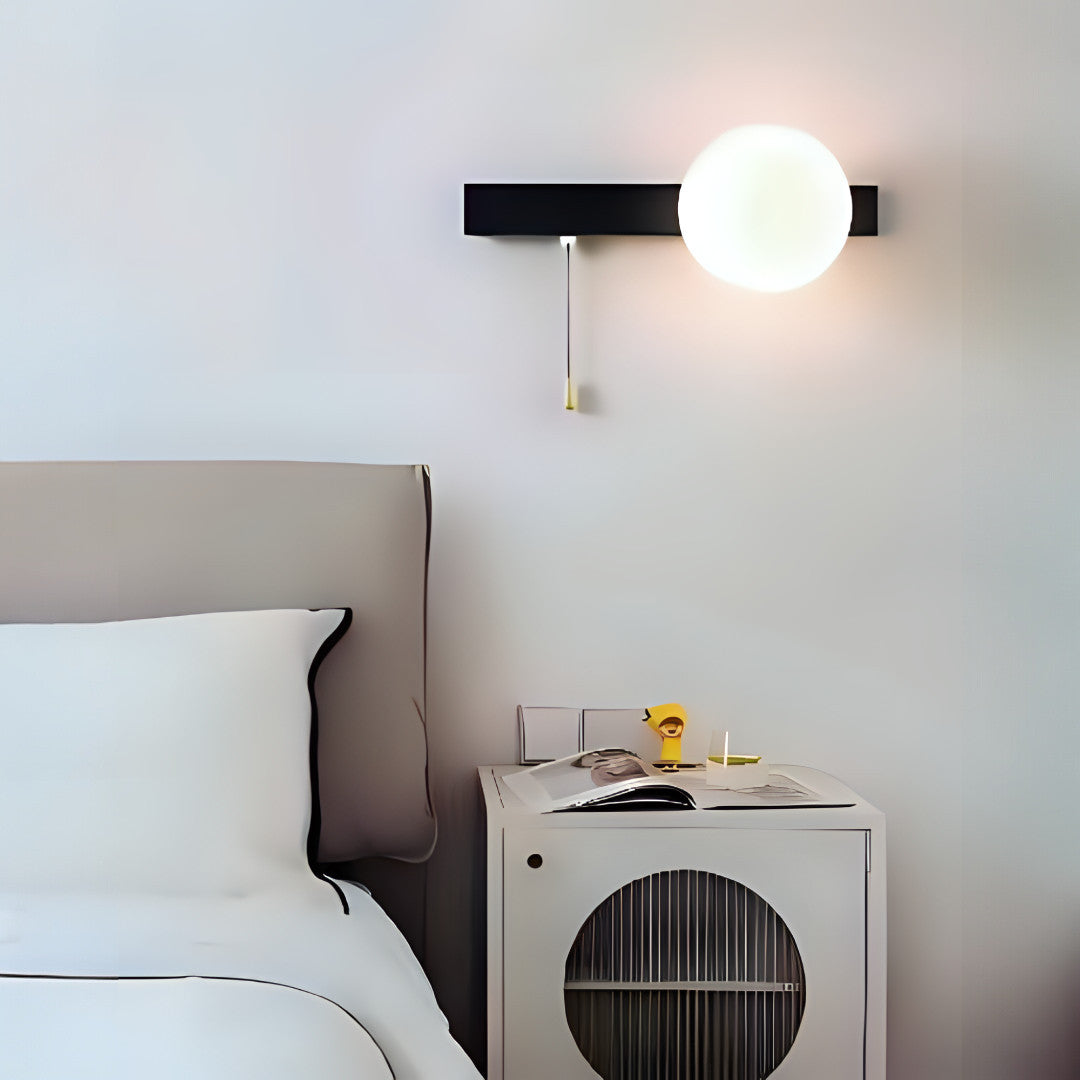 A bedroom wall light with a black horizontal metal bar and a glowing white globe, mounted beside a bed, featuring a gold pull chain switch for easy operation.
