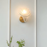 A decorative seashell-shaped wall sconce with a gold circular base and a sculpted white glass shell adorned with pearl-like accents, emitting a warm glow.
