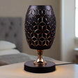A black table lamp with a geometric design, gold accents, and a sleek base sits on a wooden table. It features an LED light with an E27 bulb, casting a warm, stylish glow.