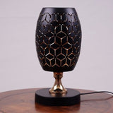 A black table lamp with a geometric design, gold accents, and a sleek base sits on a wooden table. It features an LED light with an E27 bulb, casting a warm, stylish glow.