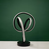 a contemporary black  table lamp featuring a curved metallic frame with an integrated circular LED light. it is placed on a round white table against a green background.