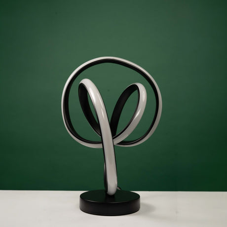 a contemporary black  table lamp featuring a curved metallic frame with an integrated circular LED light. it is placed on a round white table against a green background.
