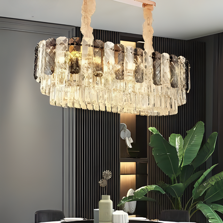 Elegant oval crystal chandelier with layered, clear and smoky glass prisms suspended from fabric-wrapped supports, casting a warm and luxurious glow in a modern dining room setting with dark paneling and lush green plants.