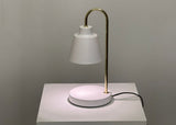 The desk lamp features a sleek and minimalist design with a white lampshade resting on a slender gold stand. The stand curves from a circular white base, creating an elegant contrast between the two tones. The lamp, equipped with an LED spotlight, emits a soft, diffused light. Positioned against a neutral background.