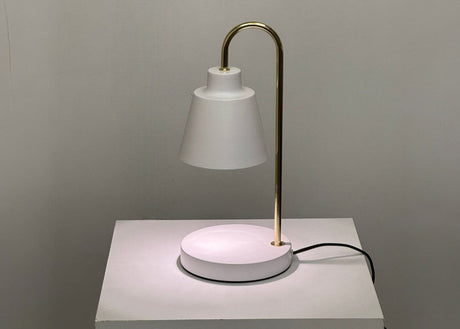 The desk lamp features a sleek and minimalist design with a white lampshade resting on a slender gold stand. The stand curves from a circular white base, creating an elegant contrast between the two tones. The lamp, equipped with an LED spotlight, emits a soft, diffused light. Positioned against a neutral background.